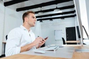 Concentrated young businessman using computer and mobile phone at workplace